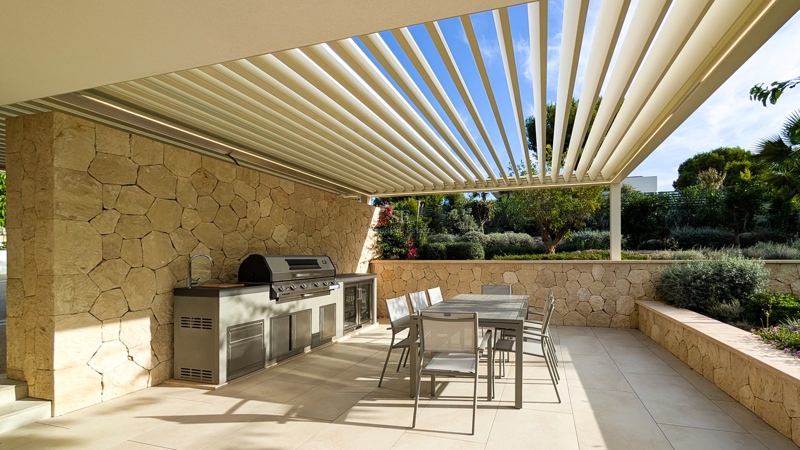 Luxury outdoor kitchen with stainless steel grill and dining table under a louvered pergola with stone walls.