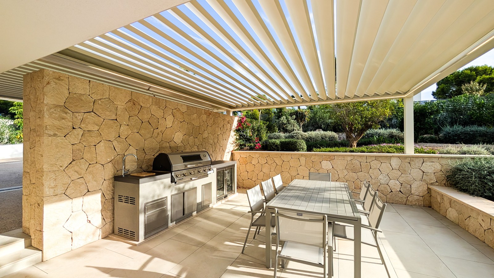 Outdoor kitchen with stainless steel grill and sink under white louvered pergola next to textured stone wall.