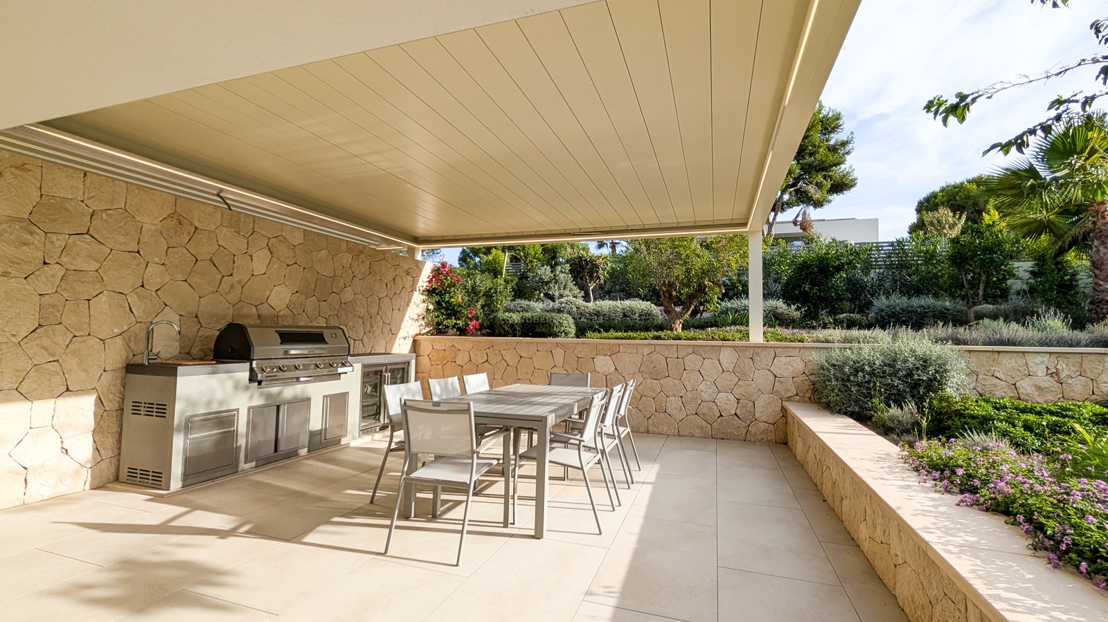 Modern outdoor kitchen with stainless steel BBQ and dining table under a pergola, surrounded by natural stone walls.