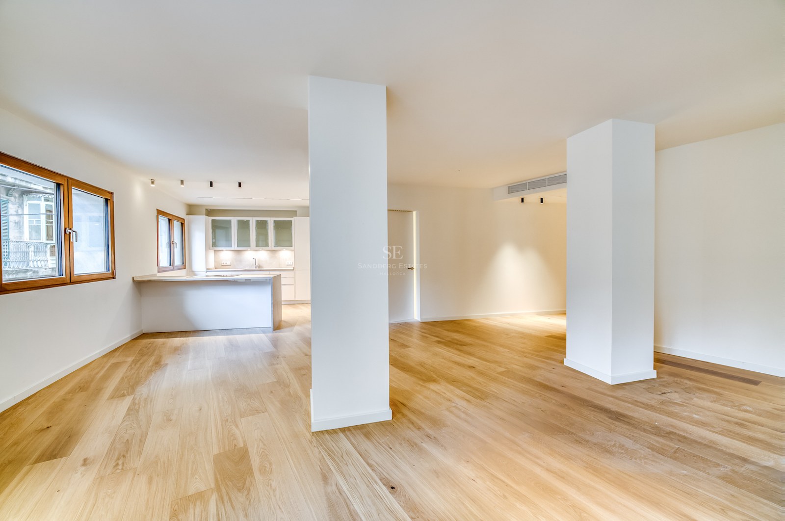 Grand espace de vie ouvert avec parquet en chêne clair, piliers blancs et cuisine minimaliste en arrière-plan.