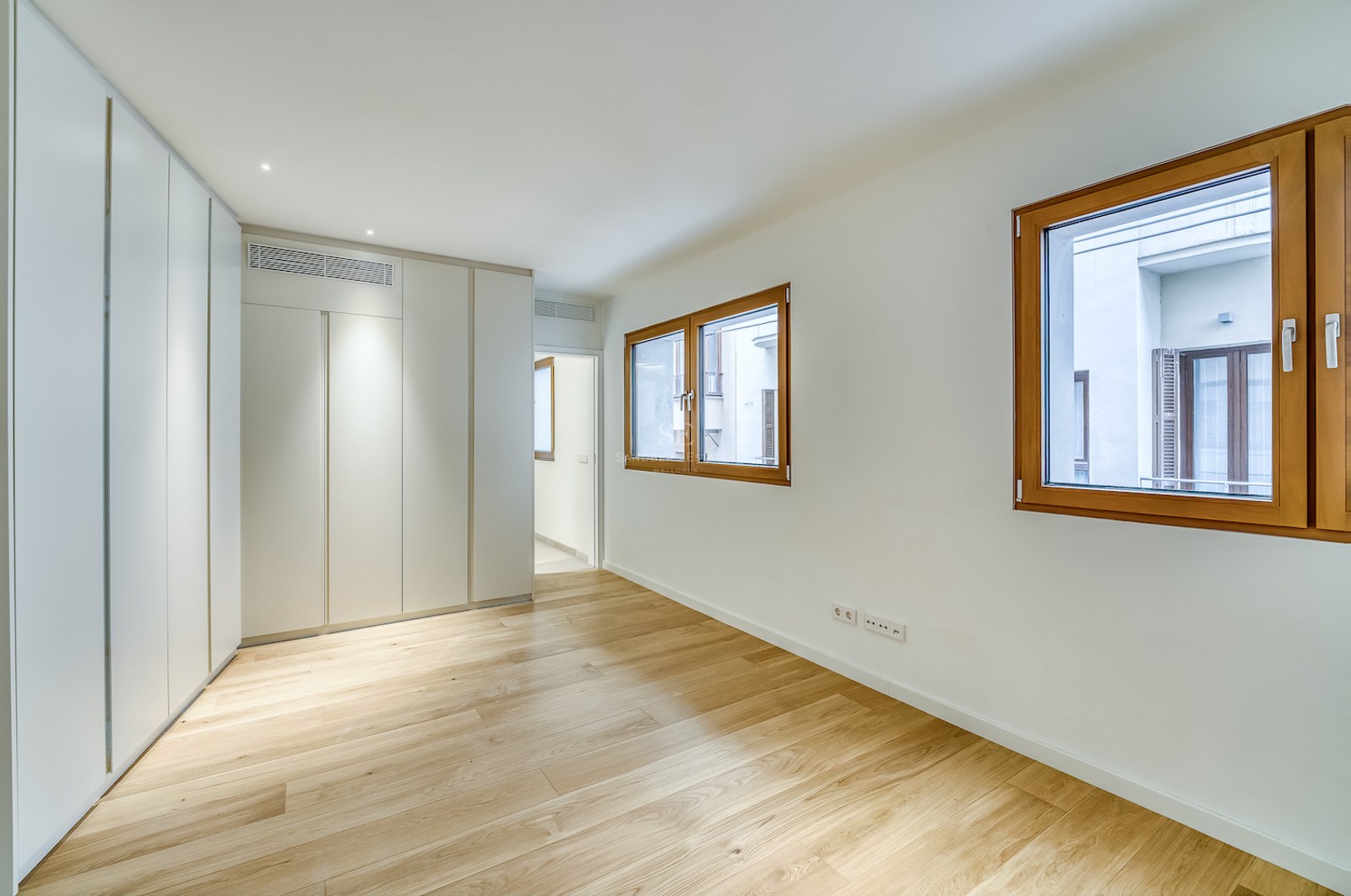 Chambre moderne vide avec parquet en chêne clair, placards blancs intégrés et deux fenêtres à cadre en bois.