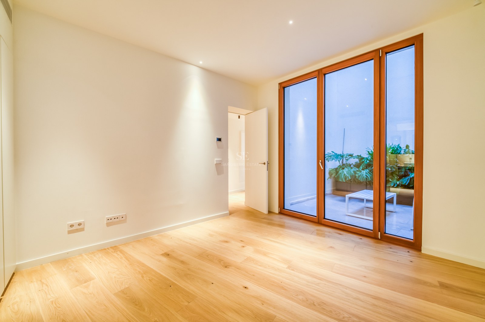 Pièce vide avec parquet en chêne, murs blancs et grandes baies vitrées en bois ouvrant sur un patio arboré.