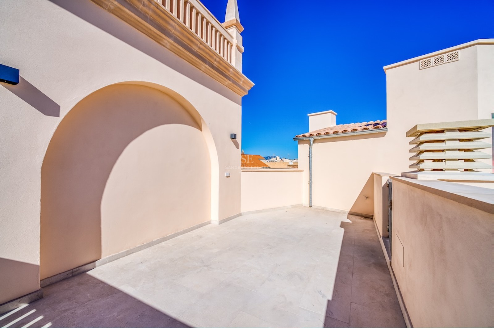 Terrasse ensoleillée sur le toit avec murs en stuc crème, sol en pierre et arche architecturale sous un ciel bleu.