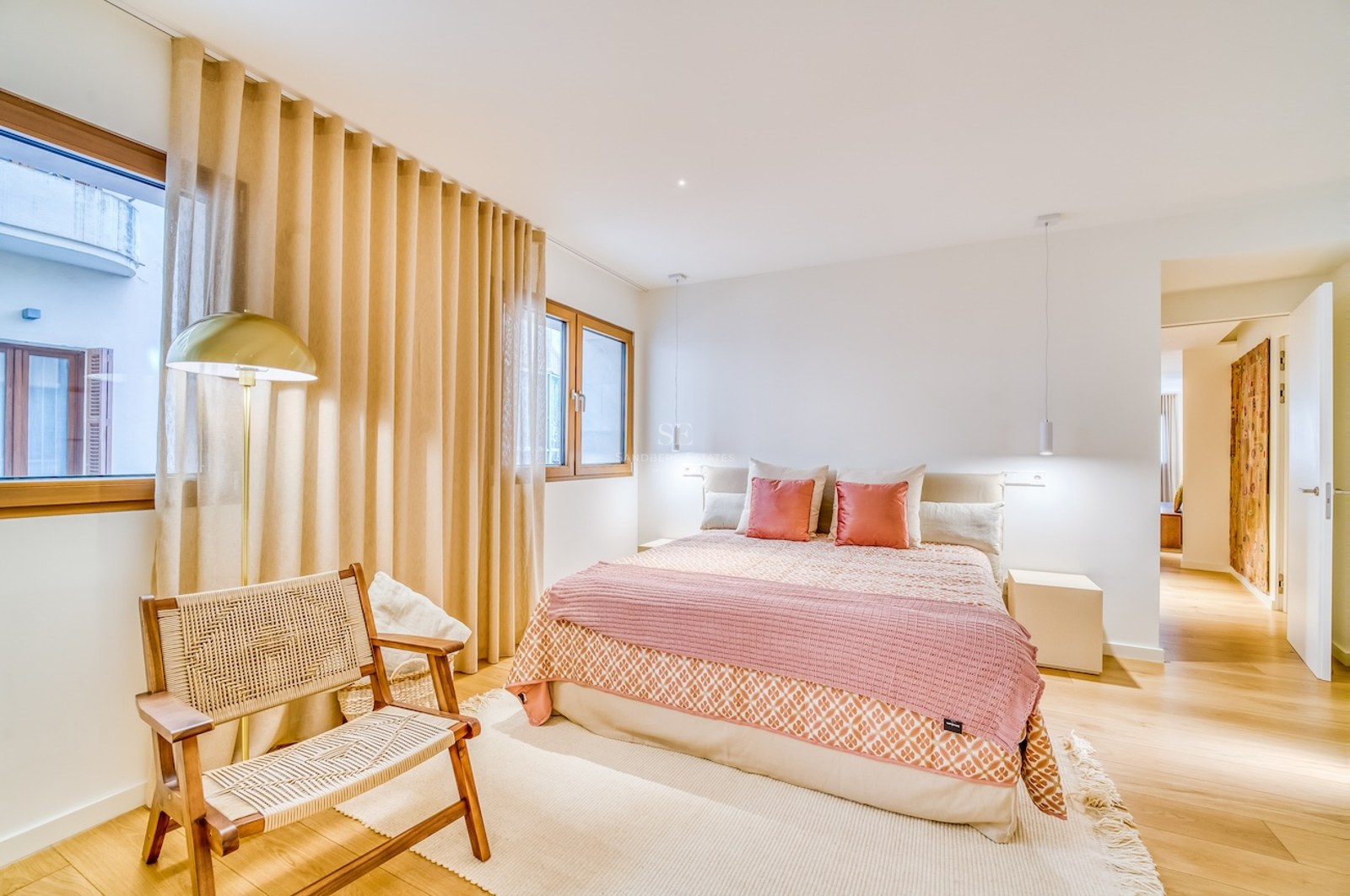 Chambre lumineuse avec lit double, textiles roses, parquet en bois, fauteuil en osier et fenêtres avec rideaux.