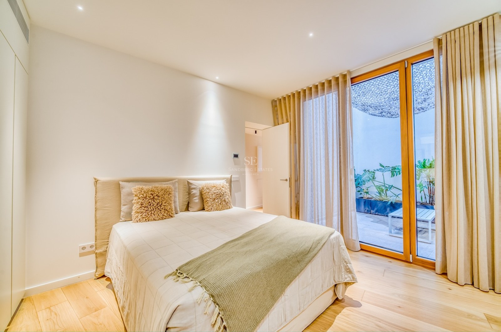 Chambre lumineuse avec parquet, literie neutre et baies vitrées ouvrant sur un patio arboré.