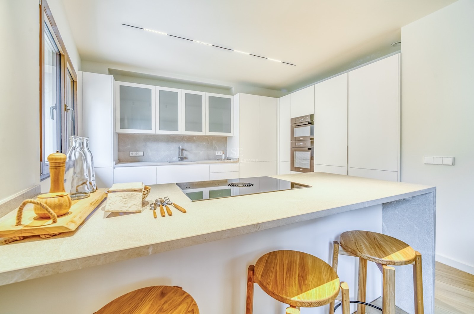 Cuisine minimaliste lumineuse avec placards blancs, îlot en pierre avec tabourets en bois et appareils intégrés.