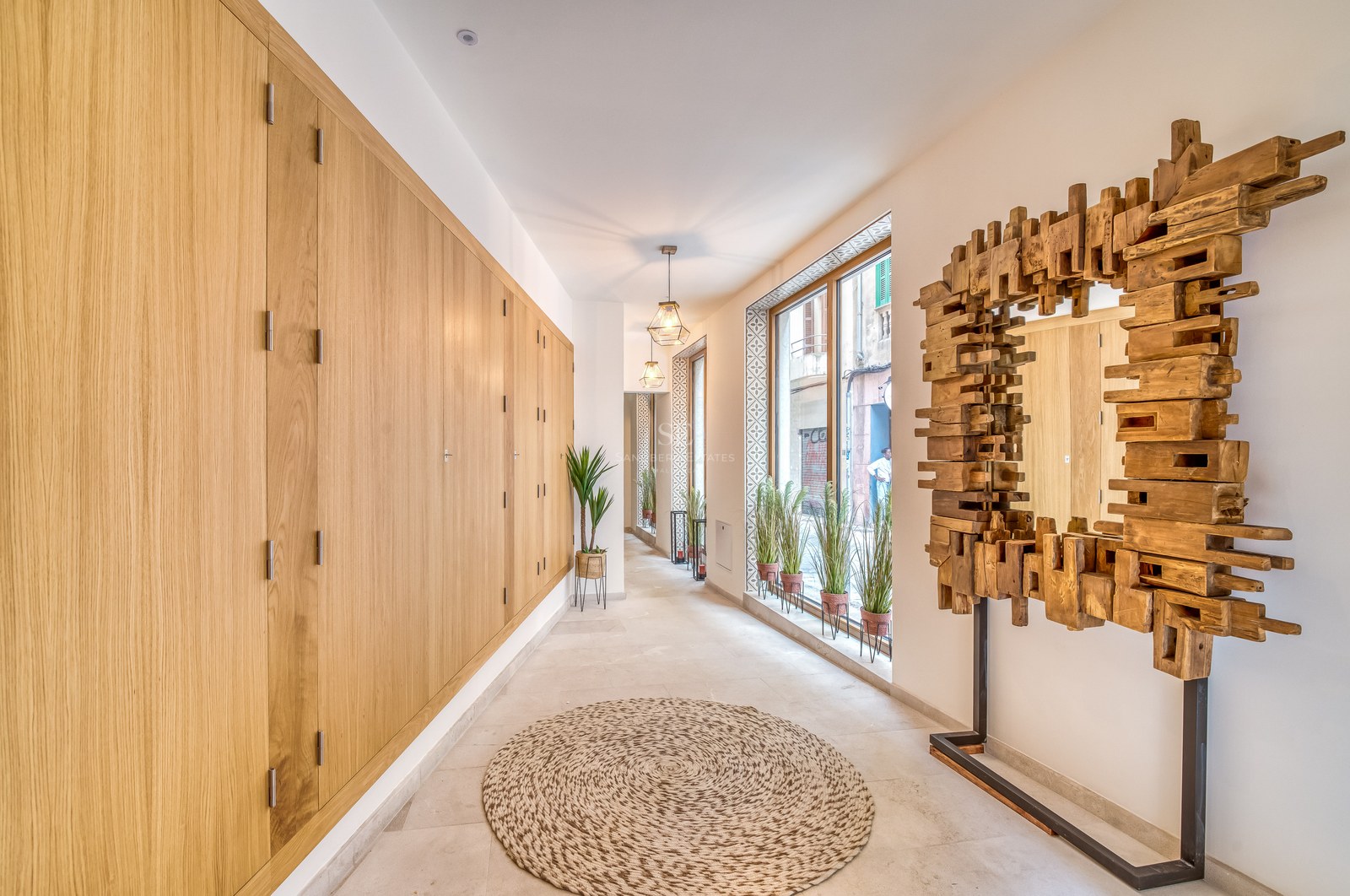 Couloir lumineux avec rangements en chêne du sol au plafond, miroir sculptural en bois et grandes fenêtres.