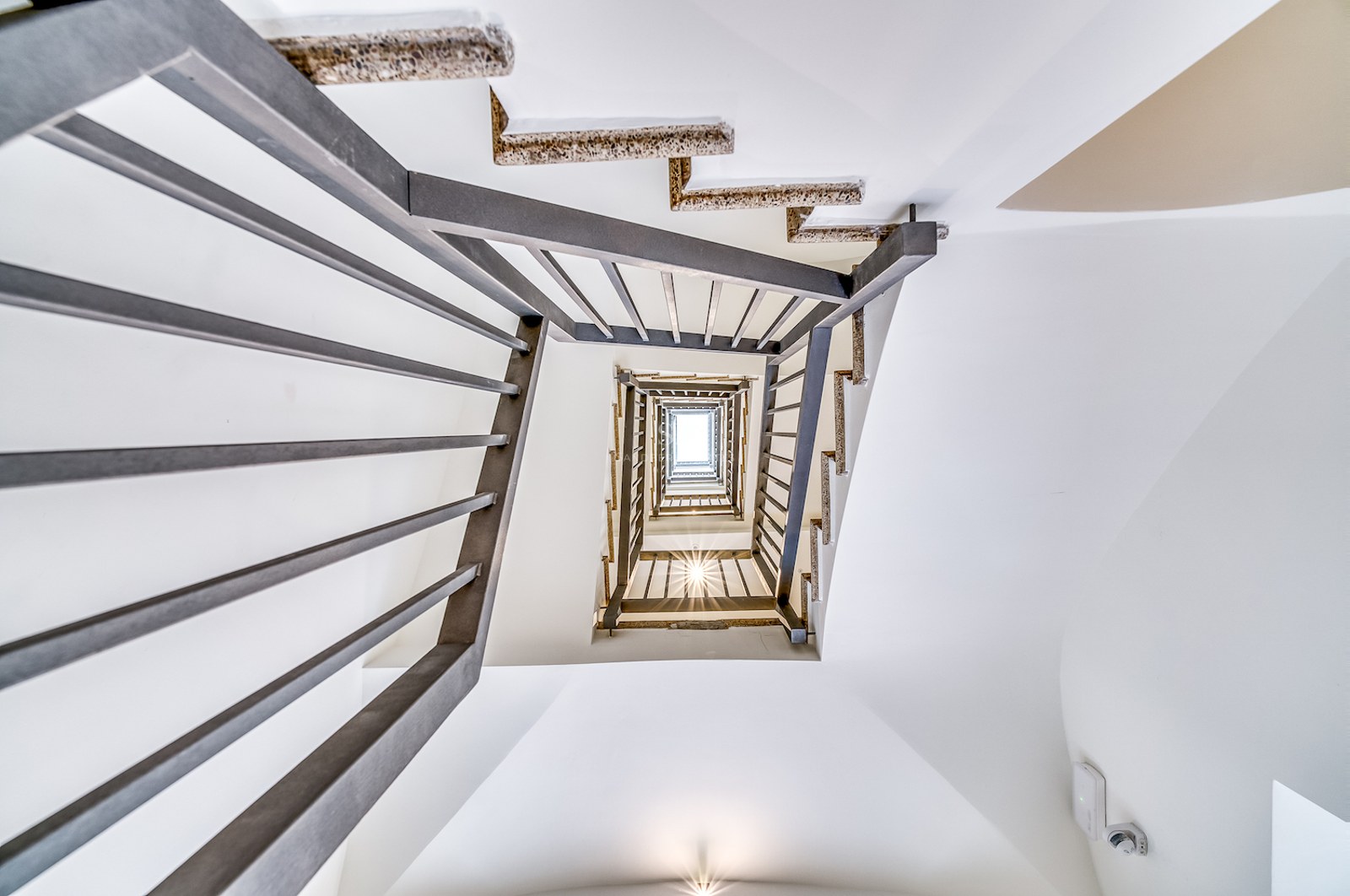 Vue vers le haut d'une cage d'escalier carrée avec rampes en métal sombre et marches en pierre sur fond de murs blancs.