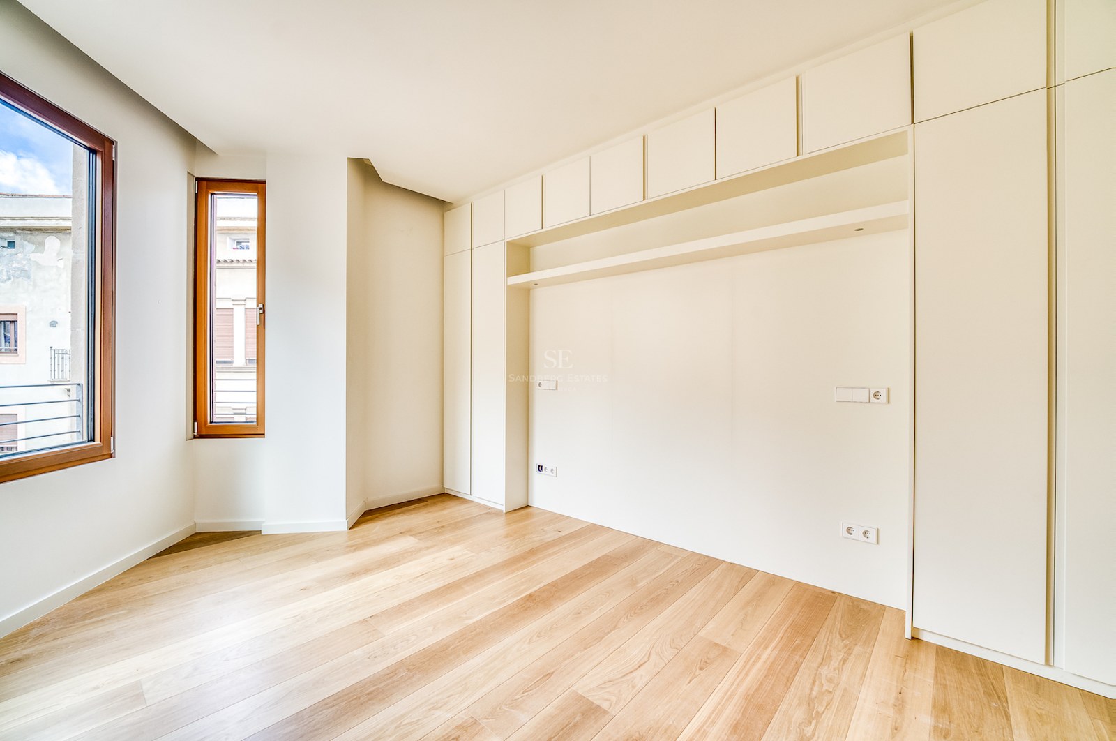 Chambre moderne vide avec placards crème toute hauteur, parquet en chêne clair et fenêtres en bois.