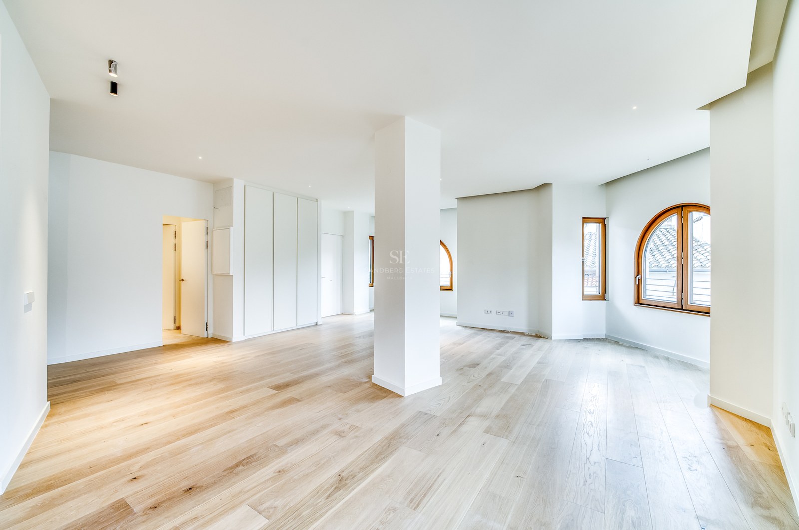 Grande pièce vide avec parquet en chêne clair, murs blancs, pilier central et fenêtres cintrées en bois.