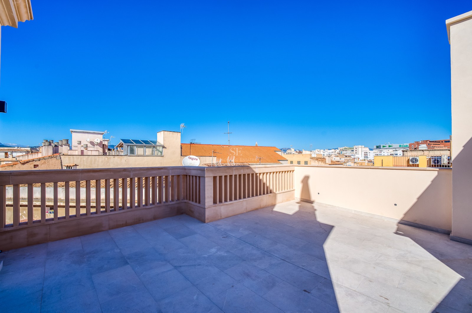 Spacious rooftop terrace with beige stone tiling and a classic balustrade overlooking a Mediterranean city under a blue sky.