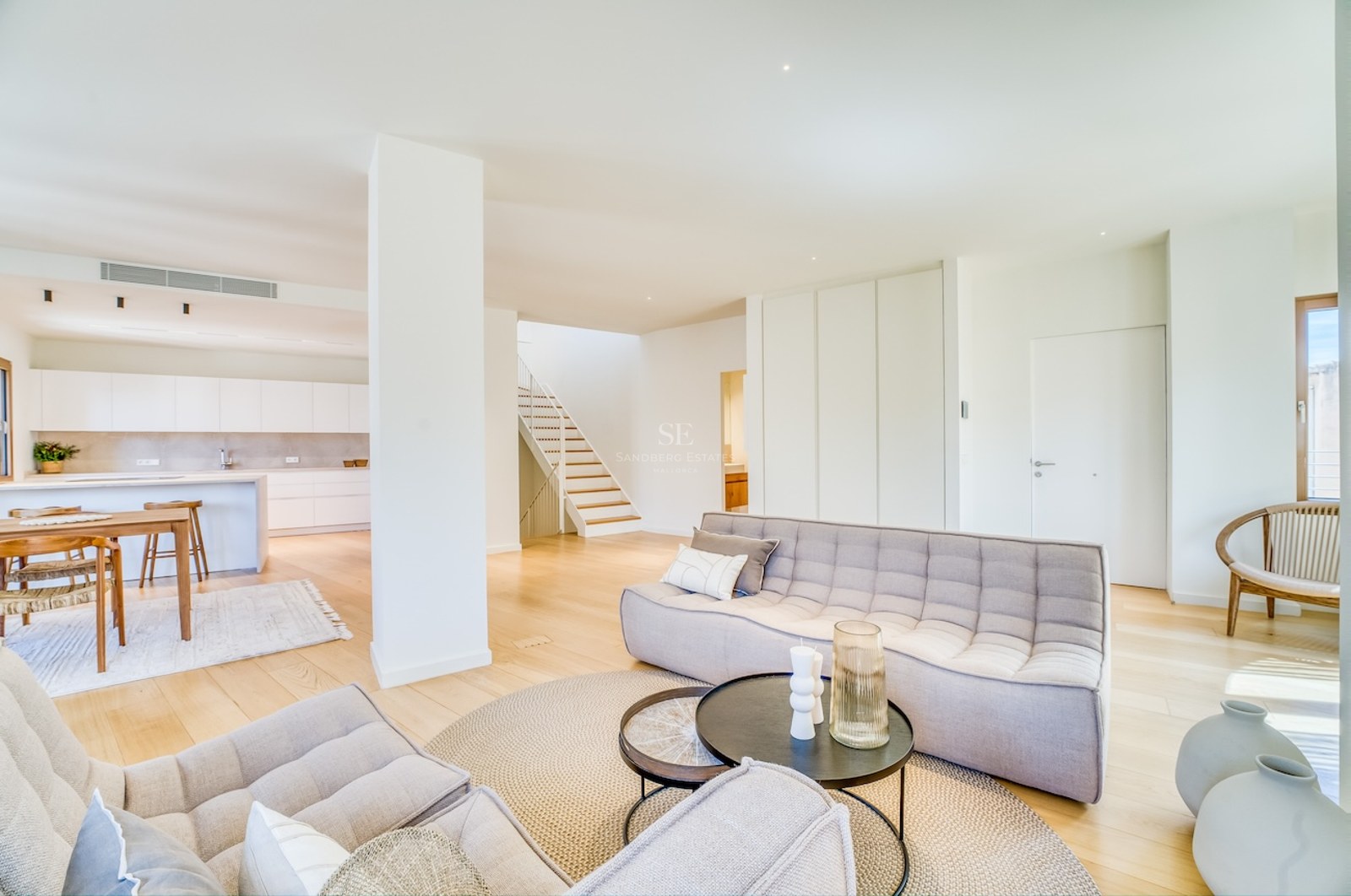 Modern open-plan living room featuring tufted grey sofas, light oak flooring, and a kitchen in the background.