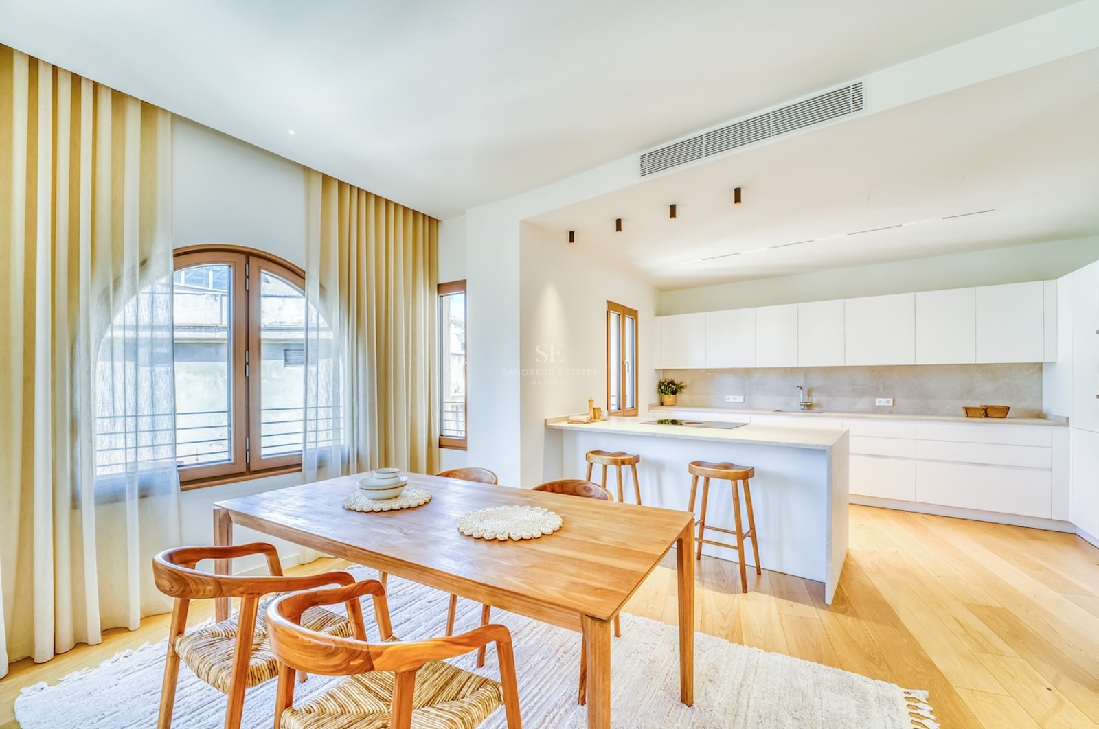 Bright open-plan kitchen with white cabinetry and a wooden dining table under an arched window with light curtains.
