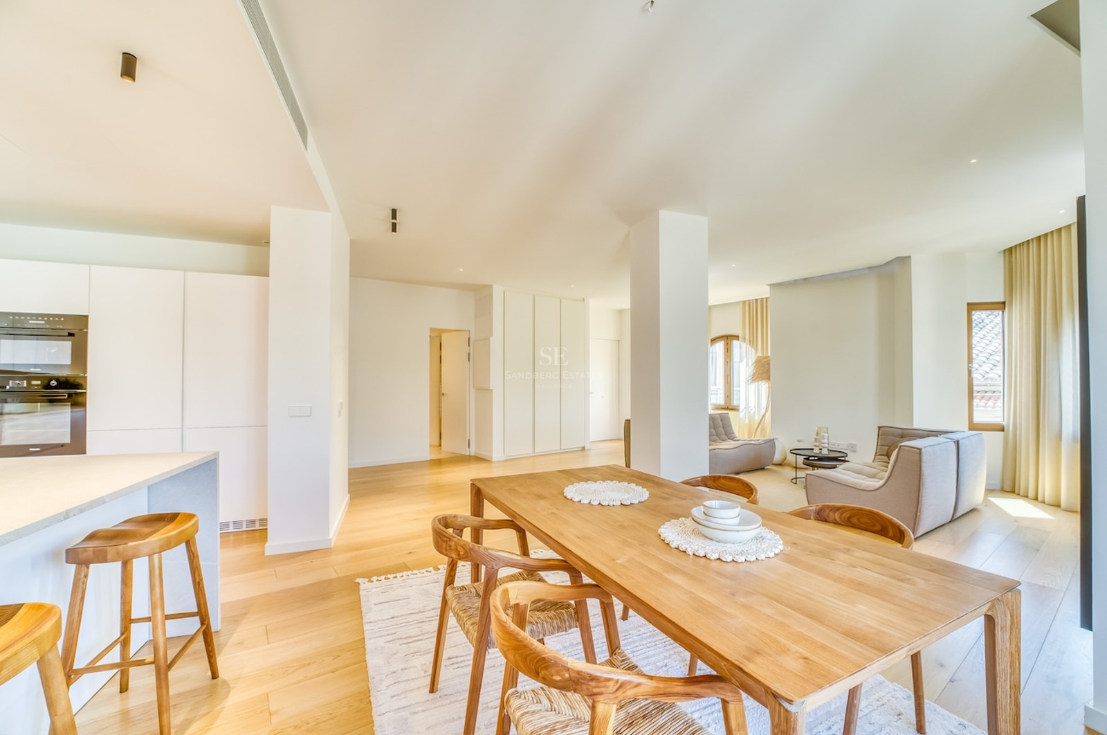 Bright open-plan living room featuring a light oak dining set, white minimalist kitchen, and floor-to-ceiling windows.