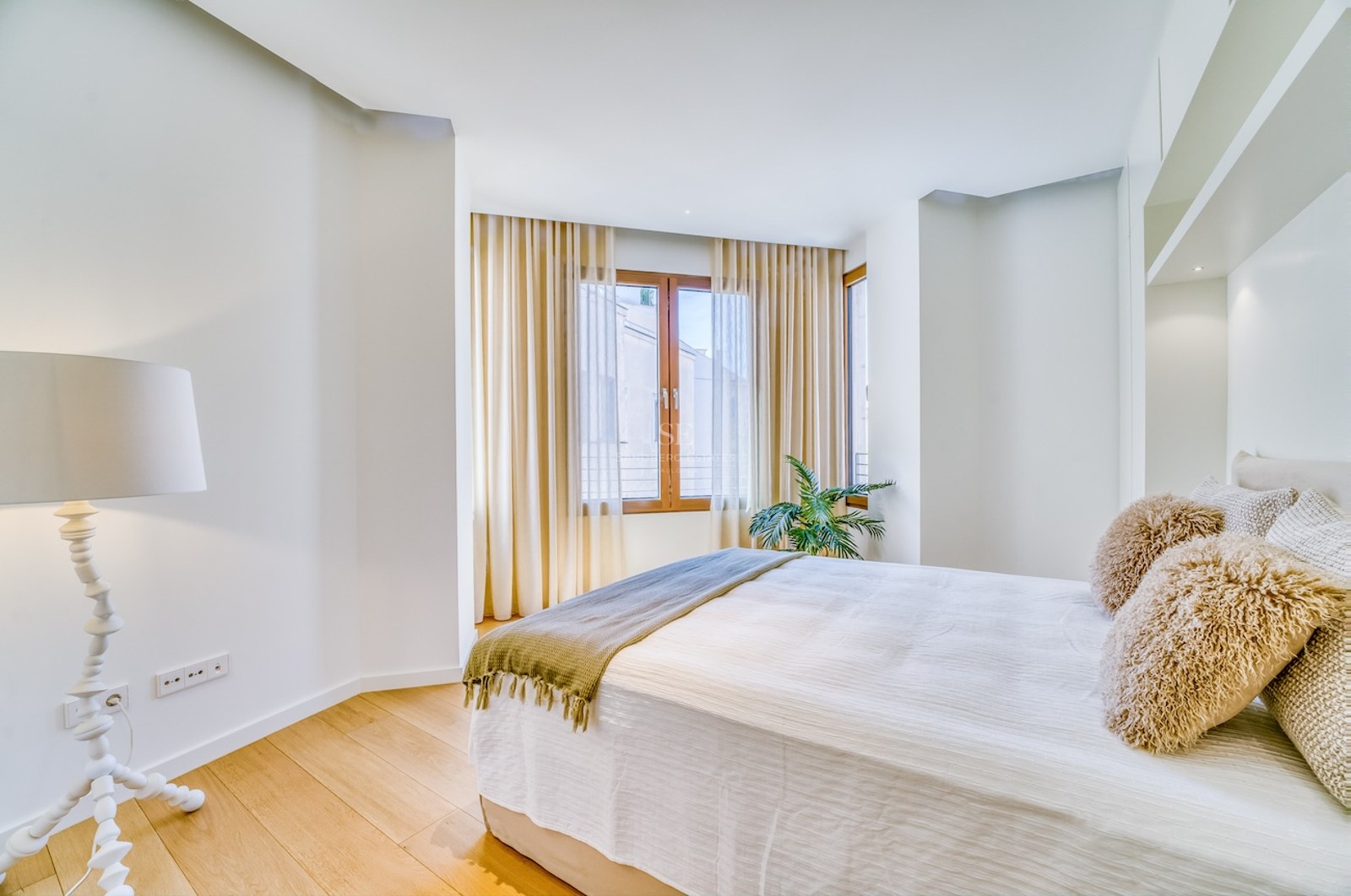 A bright bedroom with light oak floors, a double bed with textured pillows, and a large window with sheer curtains.