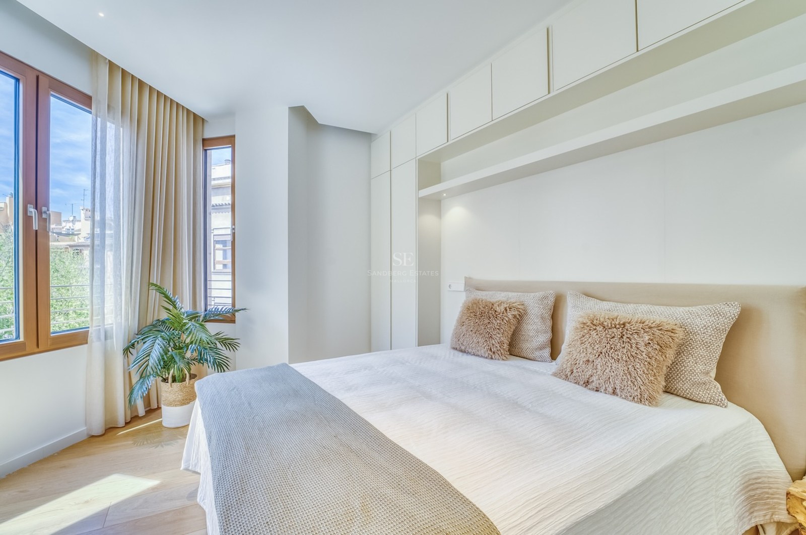 Bright master bedroom featuring integrated white storage, wooden window frames, and neutral textiles.