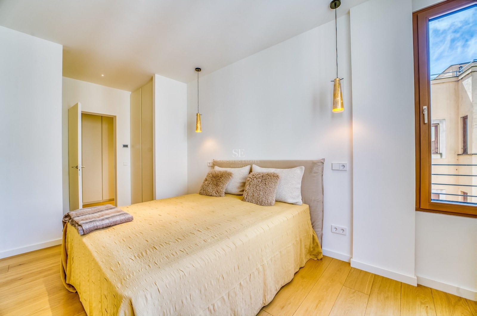 Bright bedroom featuring light wood floors, a bed with a yellow cover, and two gold pendant lamps by a window.