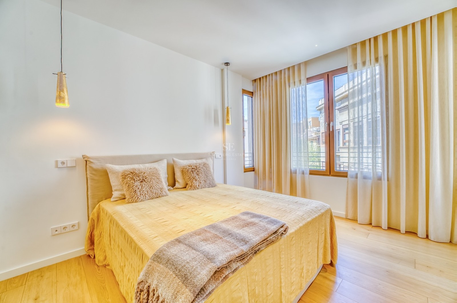 Bedroom with double bed, light wood floors, large windows, and beige curtains in a bright setting.