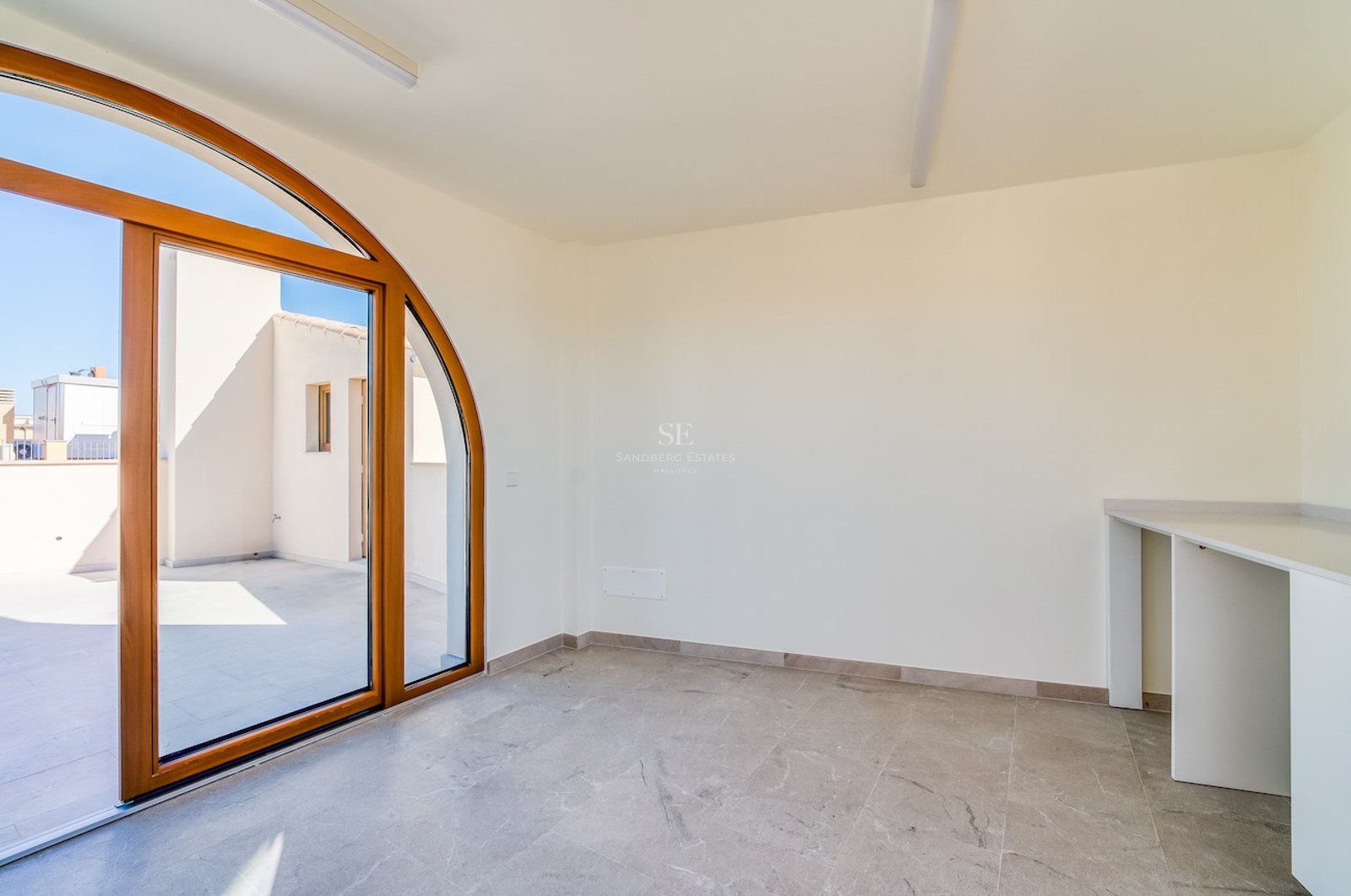 Pièce lumineuse avec sol en pierre grise, murs blancs, bureau intégré et porte arquée en bois menant à la terrasse.