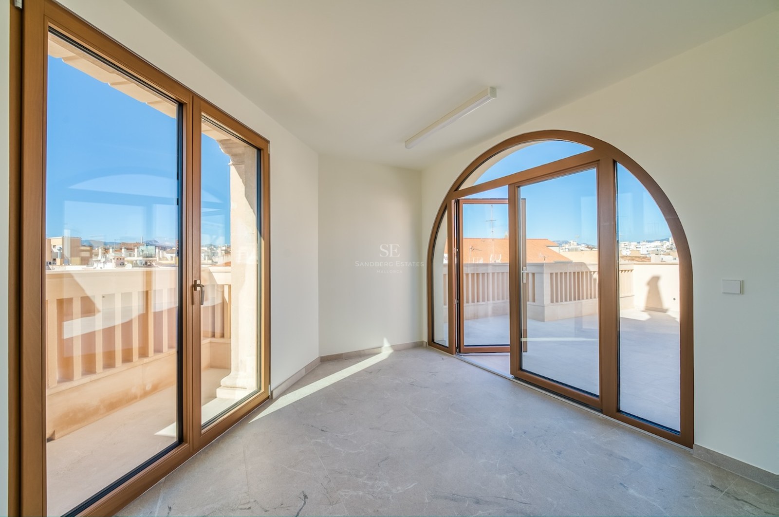 Empty room with marble floors and a large arched glass door opening to a sunny urban terrace under a clear blue sky.