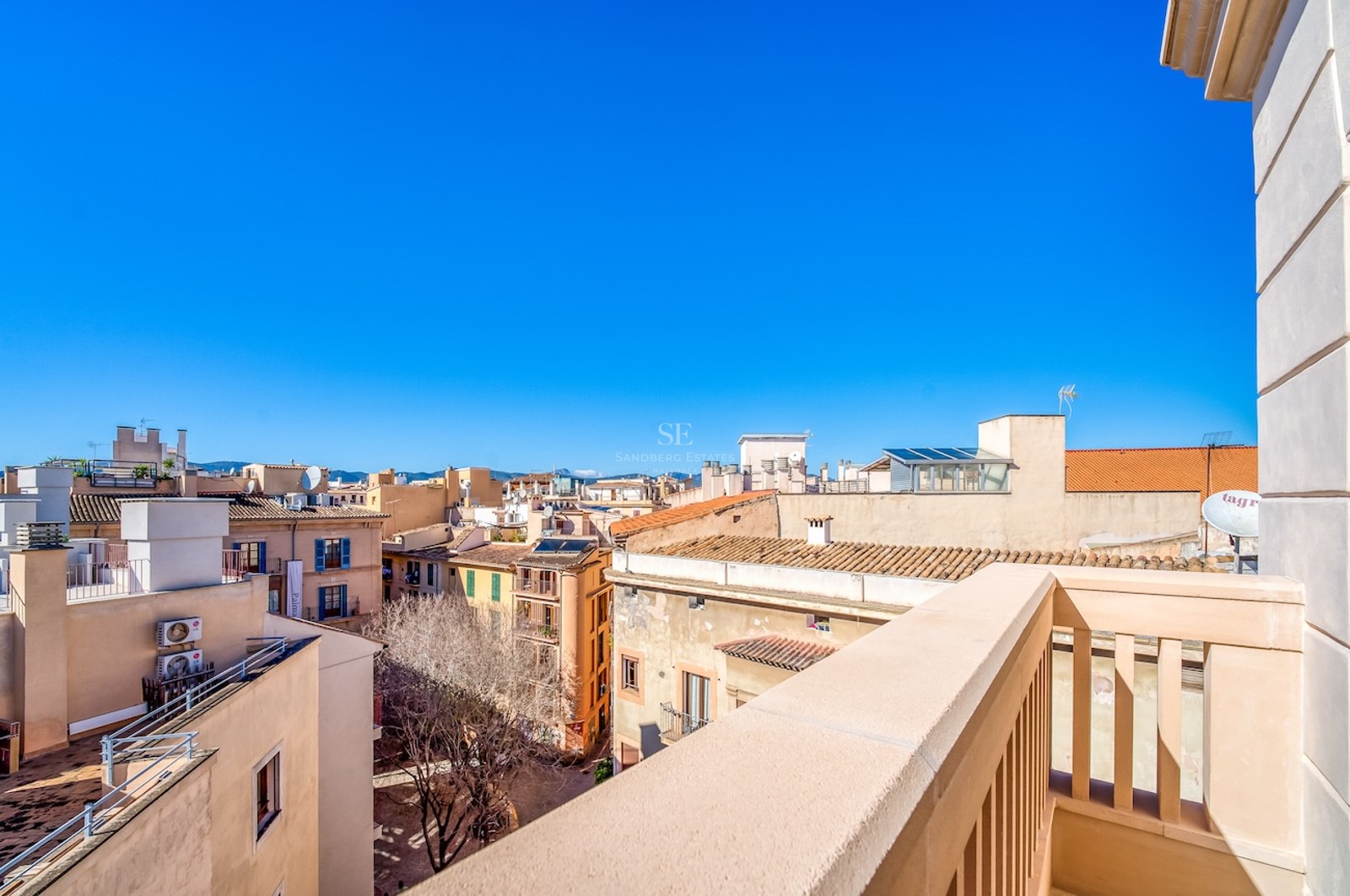 Vue surélevée depuis un balcon donnant sur les toits historiques et les montagnes lointaines sous un ciel bleu.