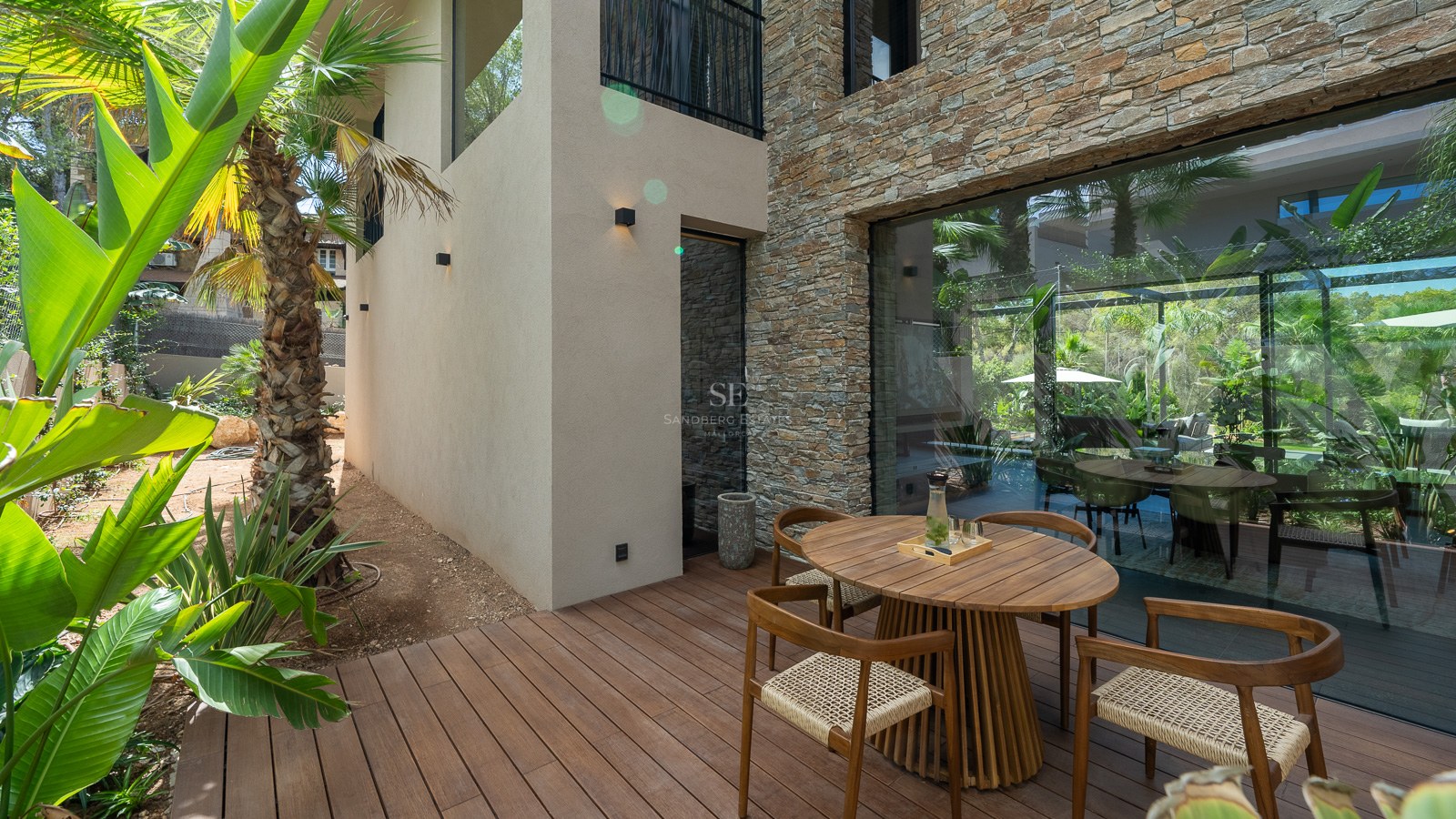 Contemporary wooden terrace featuring a round dining set, natural stone wall, and tropical garden backdrop.