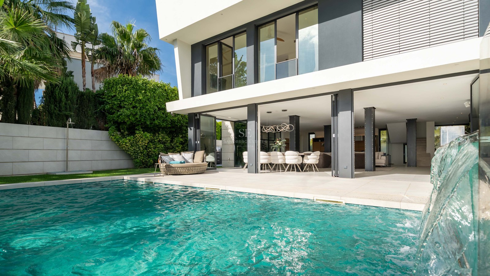 Extérieur de villa moderne avec piscine turquoise, cascade et salon ouvert sous un ciel bleu éclatant.