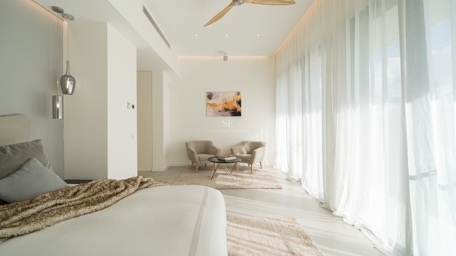 Grande chambre blanche avec baies vitrées, rideaux voilages, deux fauteuils et ventilateur de plafond en bois.