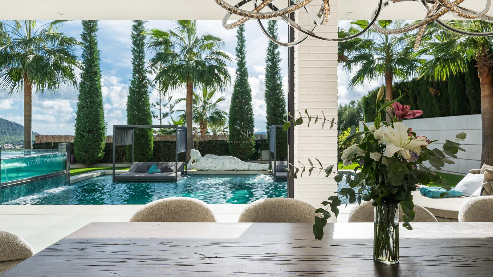 Table à manger en bois donnant sur une piscine turquoise et un jardin tropical avec palmiers et lits de jour.