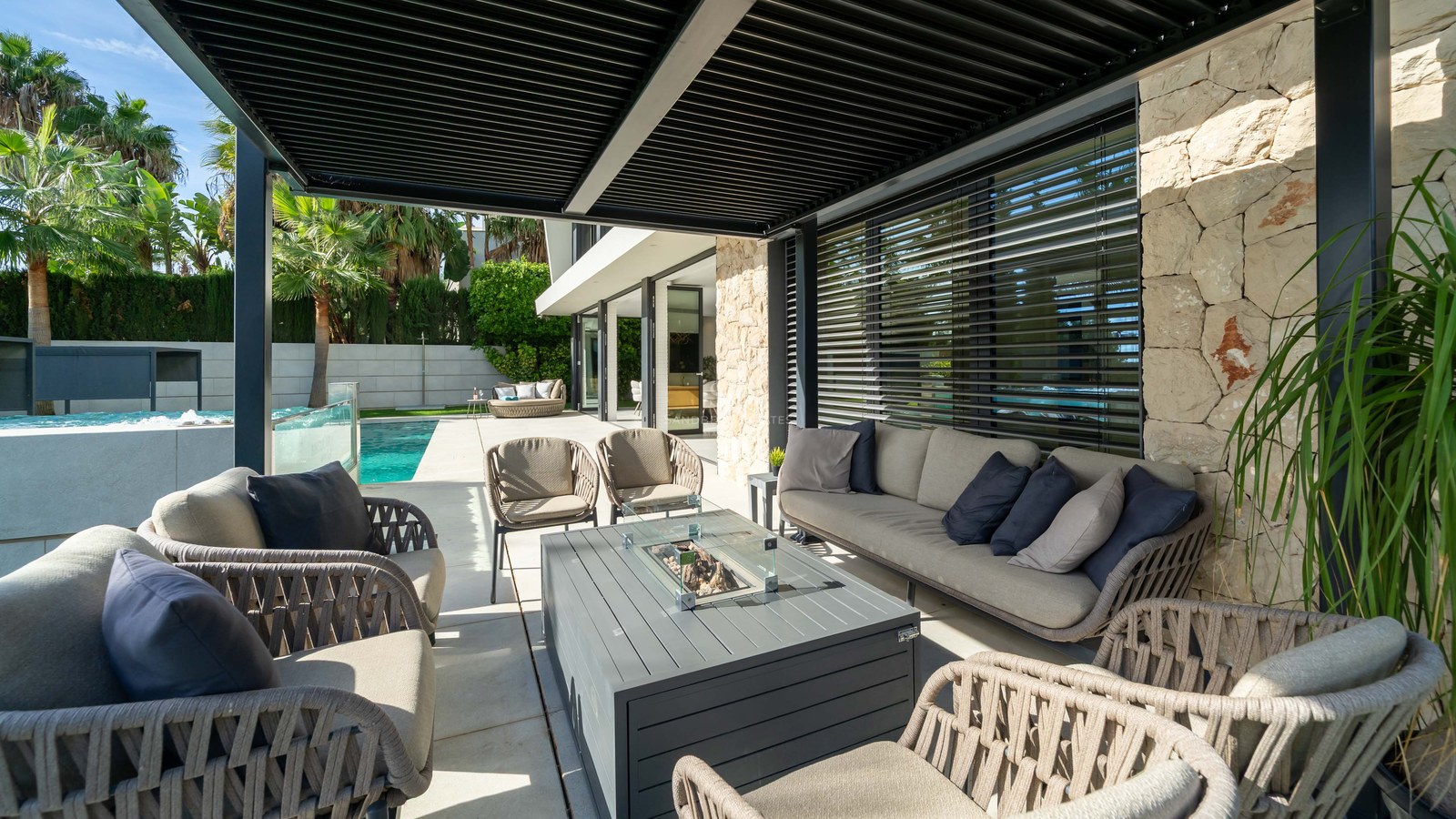 Salon extérieur de luxe avec mobilier gris sous une pergola noire, à côté d'une piscine et d'un mur en pierre.