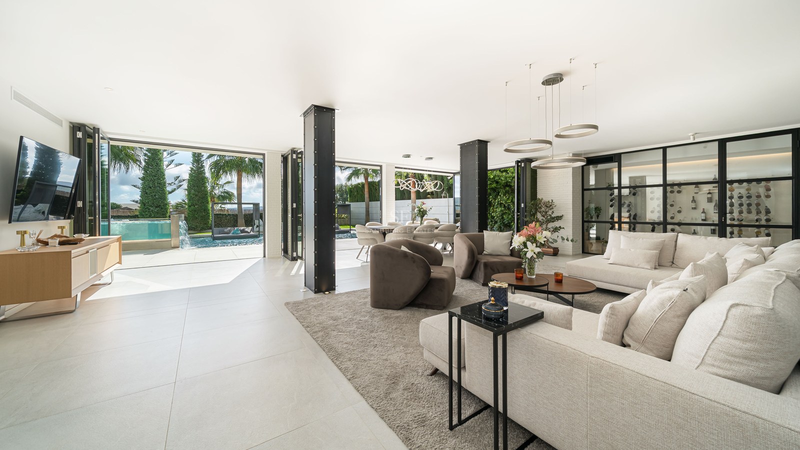 Salon moderne spacieux avec piliers en acier, canapés blancs et grandes baies vitrées ouvrant sur une piscine.