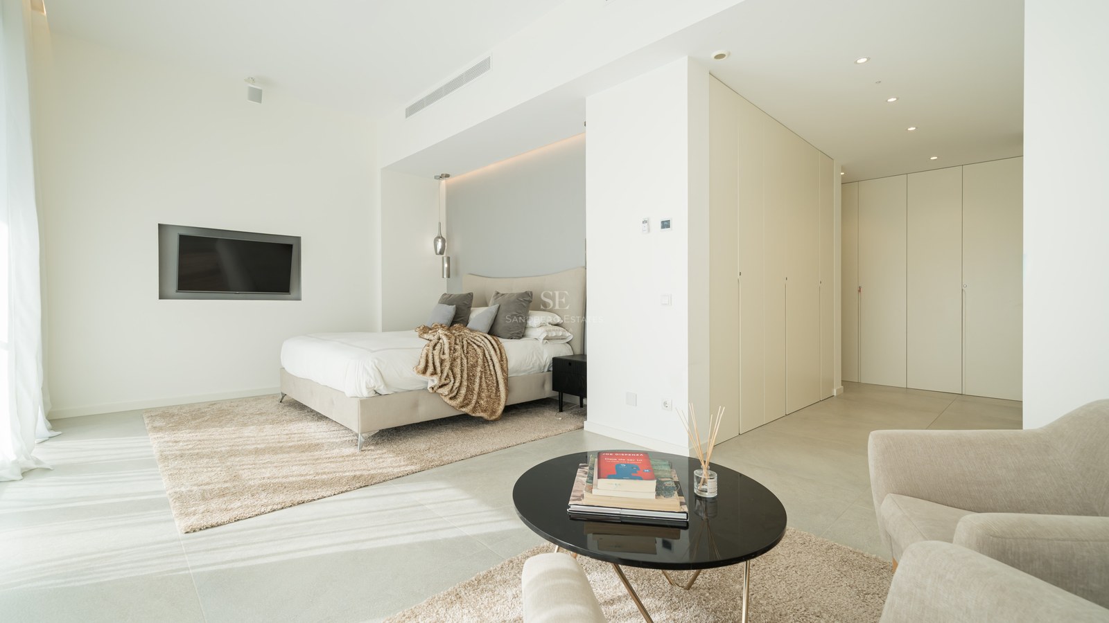 Chambre principale moderne avec lit design, placards intégrés, coin salon et sol en grès grand format.