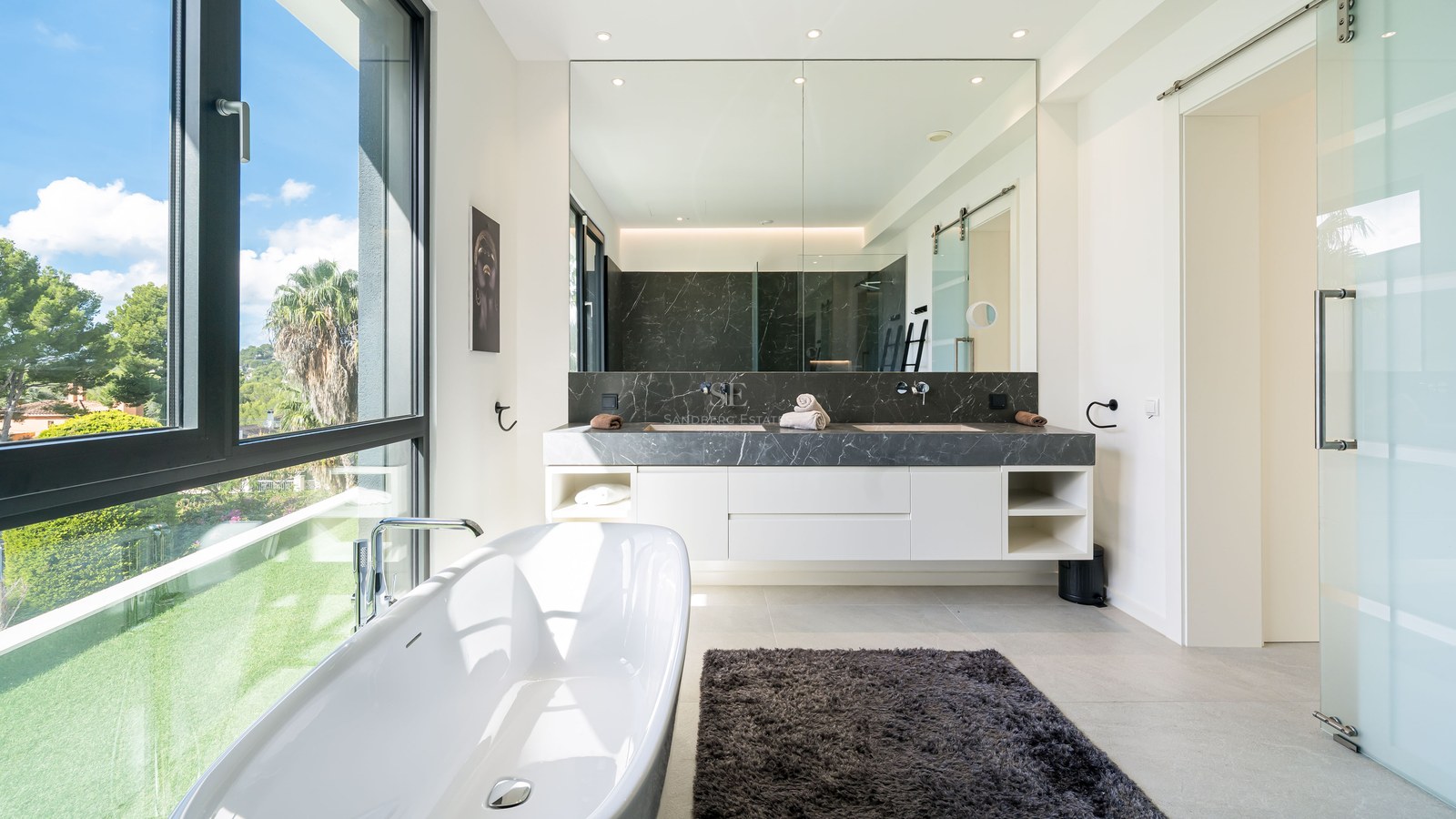 Salle de bain contemporaine lumineuse avec baignoire îlot près d'une grande fenêtre donnant sur un jardin verdoyant.