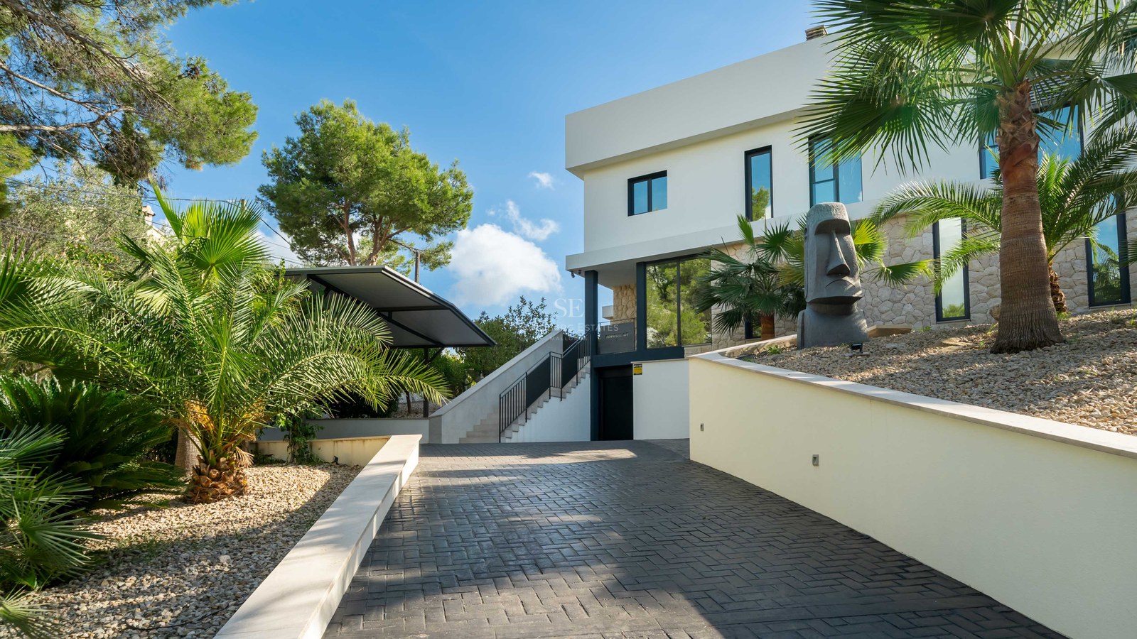 Villa blanche moderne avec accents en pierre, allée pavée, palmiers et statue Moai sous un ciel bleu.