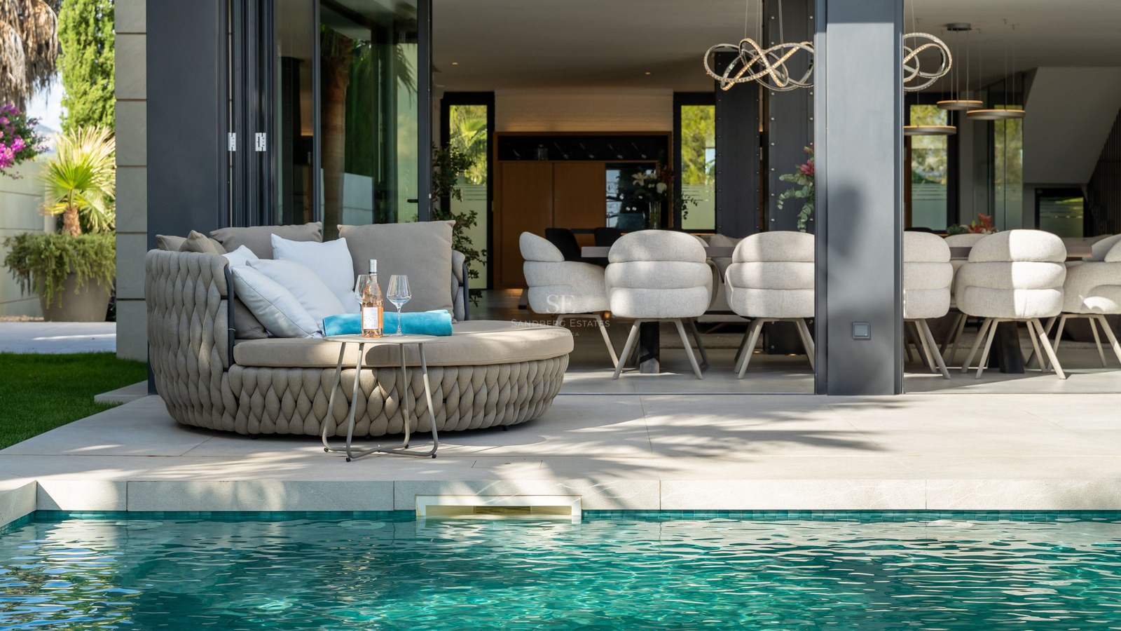 Terrasse de luxe avec bain de soleil tressé au bord d'une piscine turquoise, vue sur l'intérieur moderne.