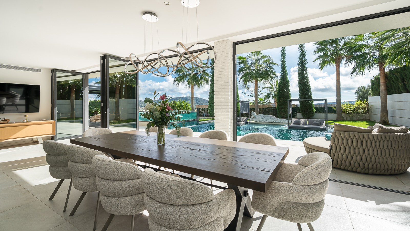Salle à manger moderne avec table en bois et chaises élégantes s'ouvrant sur une piscine privée et des palmiers.