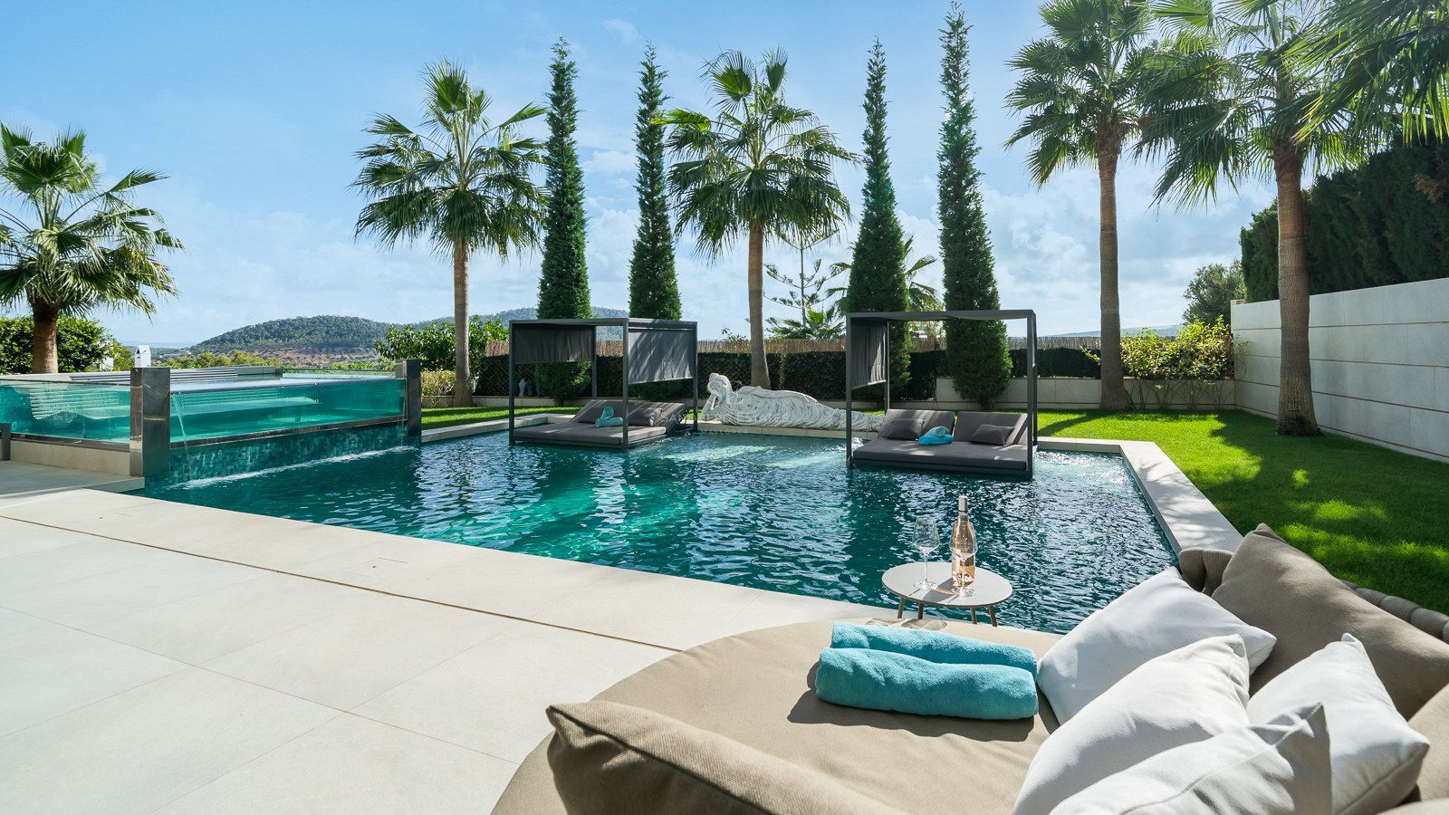 Piscine de luxe avec paroi en verre, lits intégrés, palmiers et vue sur la montagne sous un ciel bleu.
