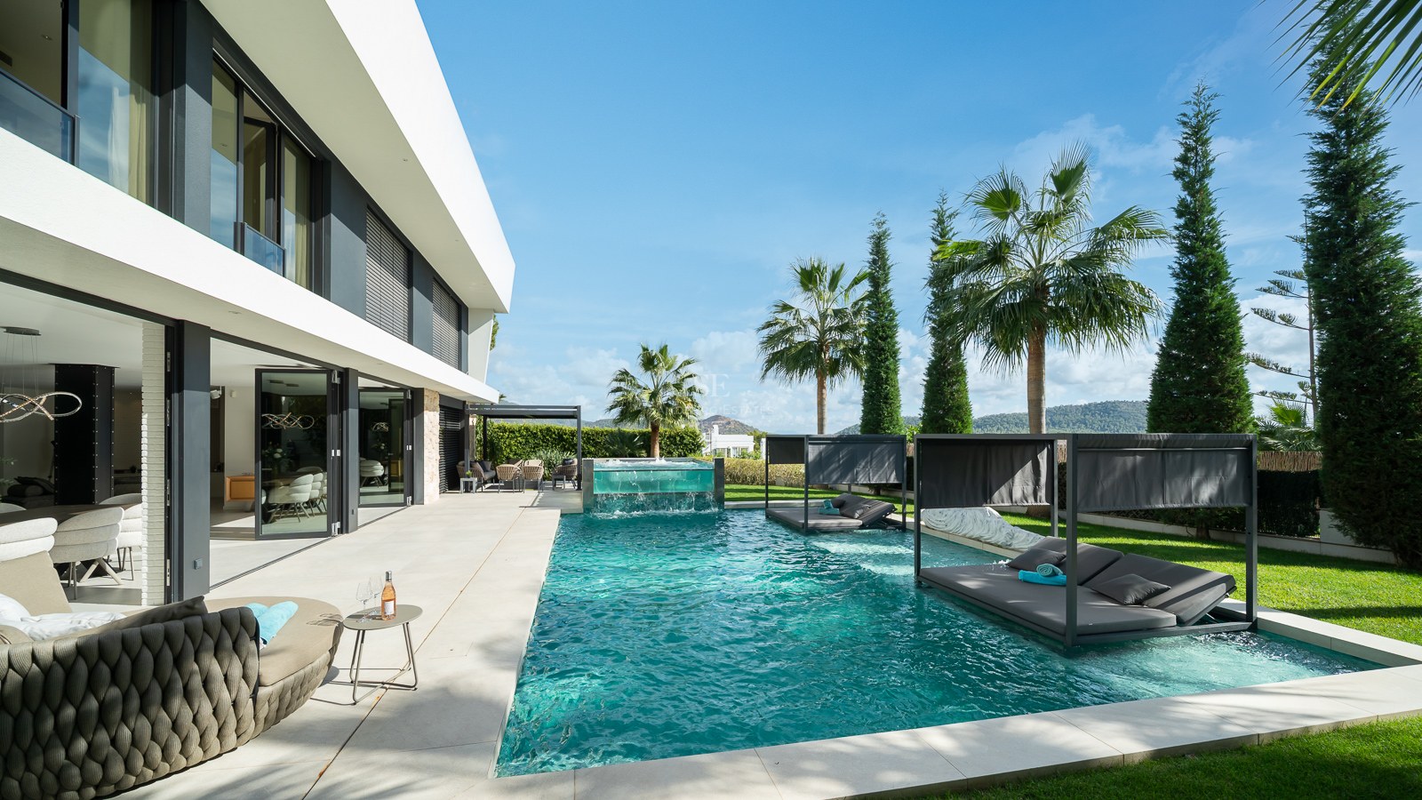 Grande piscine turquoise avec lits de repos immergés et cascade en verre devant une villa blanche moderne.