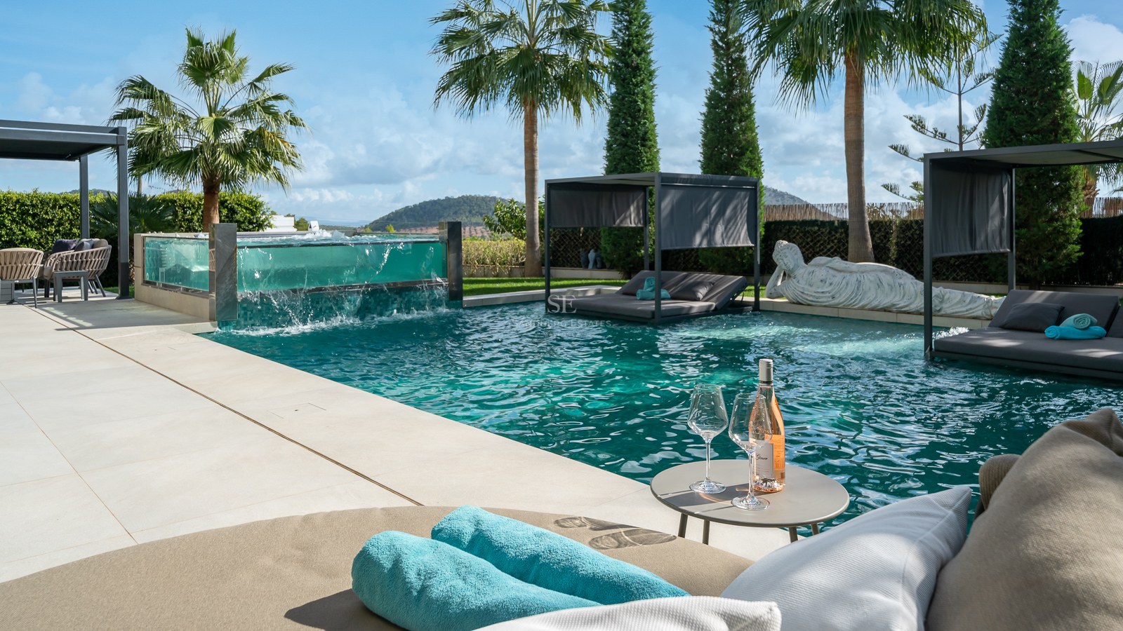Espace piscine de luxe avec spa aux parois de verre, lits de jour immergés et statue de Bouddha sous les palmiers.