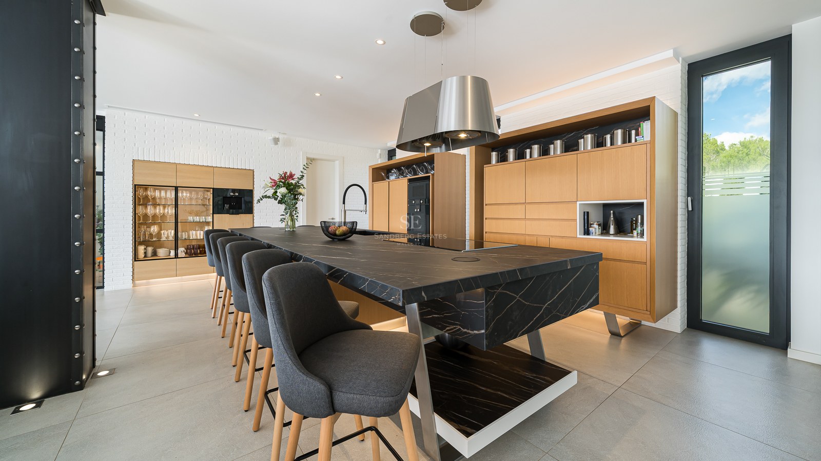 Cuisine moderne avec un grand îlot en marbre noir, tabourets gris, meubles en bois clair et mur en briques blanches.