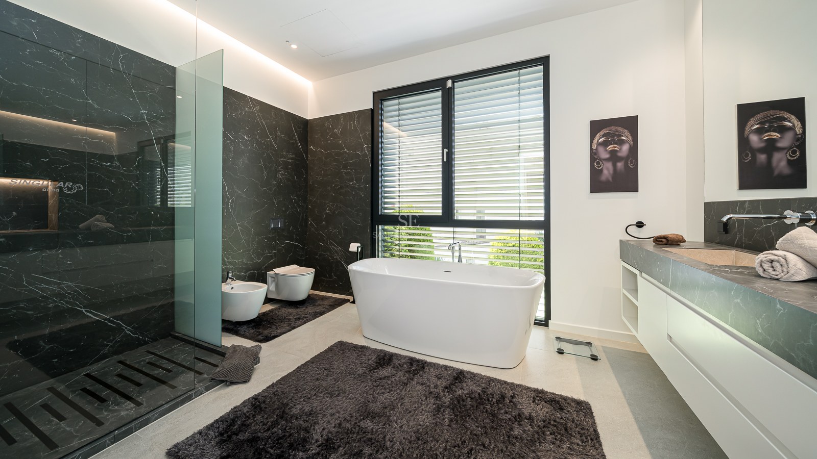 Salle de bains moderne avec baignoire blanche îlot, douche en marbre sombre et grande fenêtre avec stores.