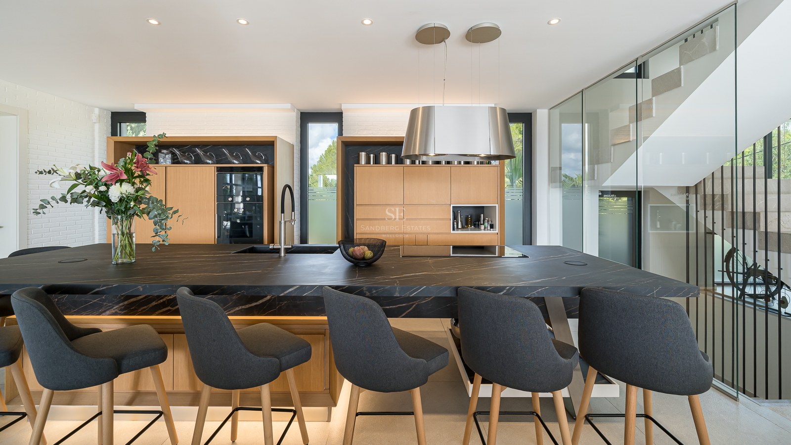 Cuisine moderne avec un grand îlot en pierre noire, tabourets gris, meubles en chêne et escalier en verre.