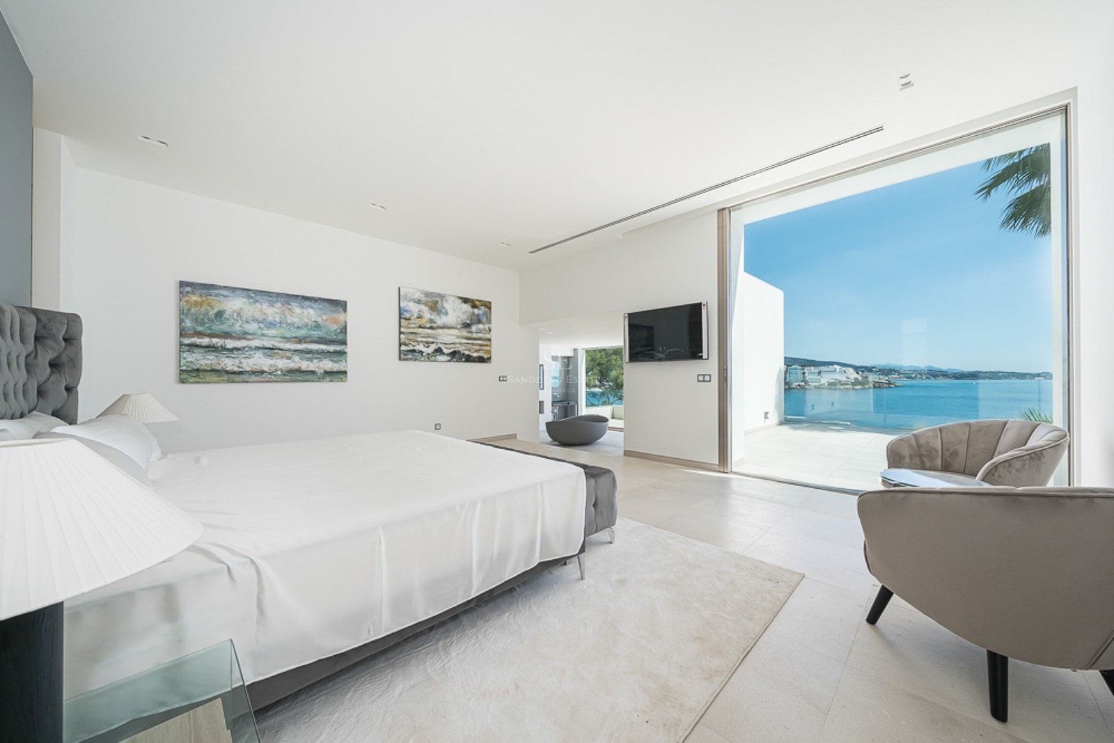 Dormitorio luminoso con cama blanca, sillones de terciopelo y grandes ventanales con vistas al mar Mediterráneo.