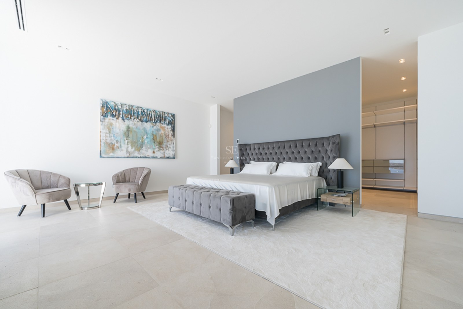 Dormitorio principal con cama capitoné gris, zona de estar de terciopelo y vestidor al fondo.