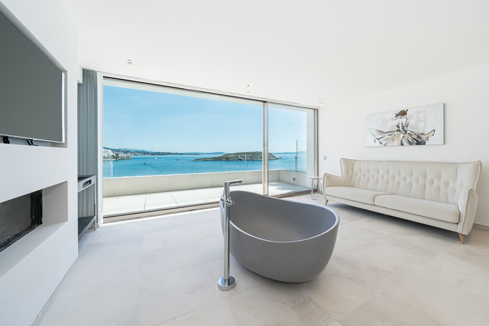 Baño moderno con bañera gris exenta, sofá crema y grandes ventanales con vistas panorámicas al mar.