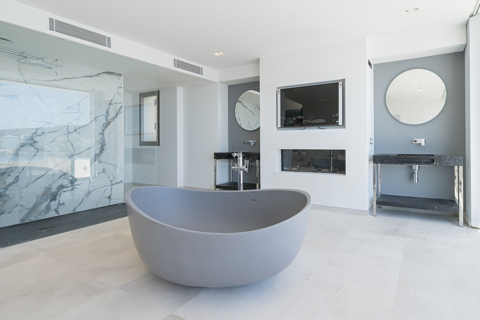 Baño moderno con bañera gris, ducha con pared de mármol y lavabos dobles con espejos redondos.