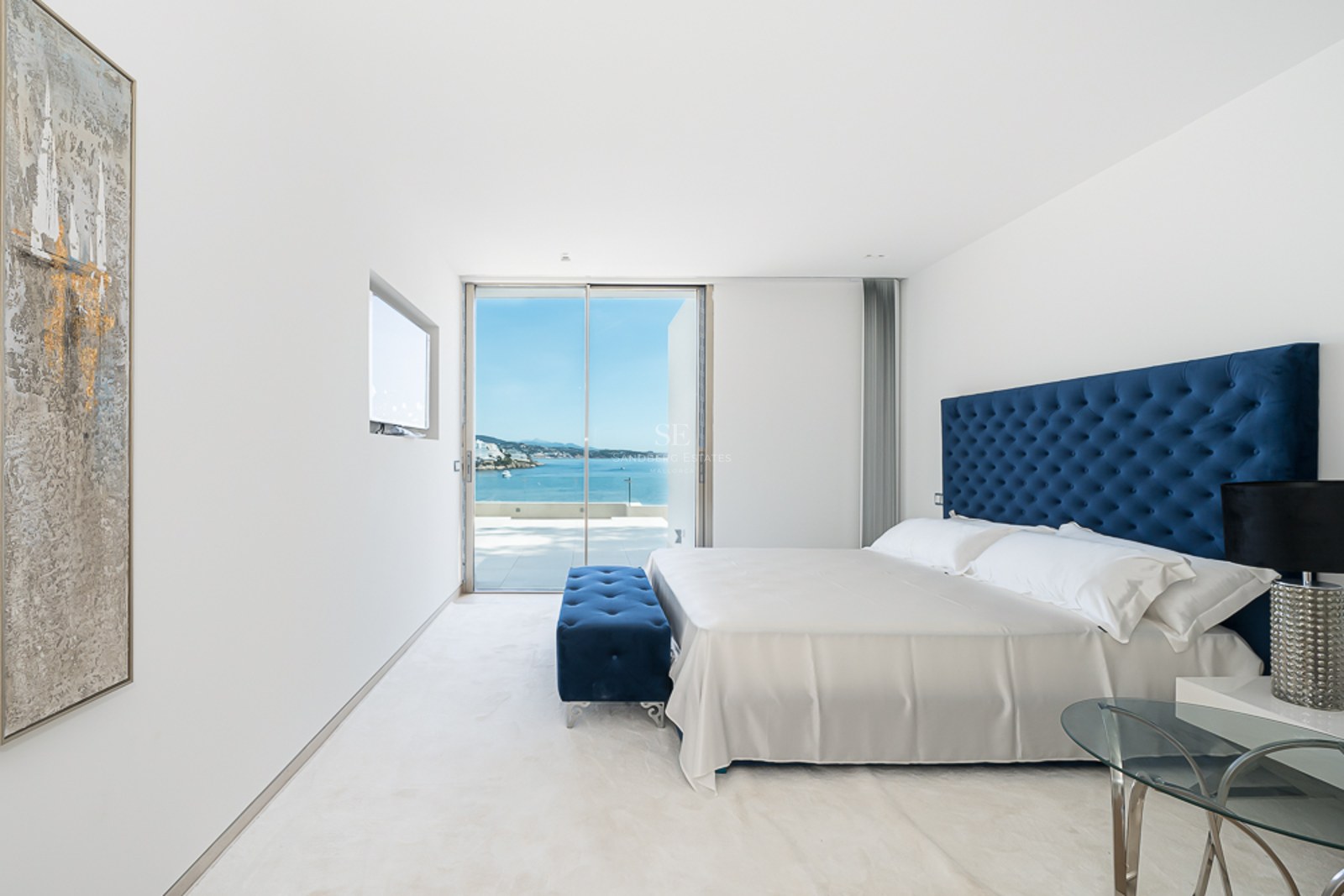 Dormitorio moderno con cabecero capitoné azul, alfombra blanca y grandes ventanales con vistas al océano.