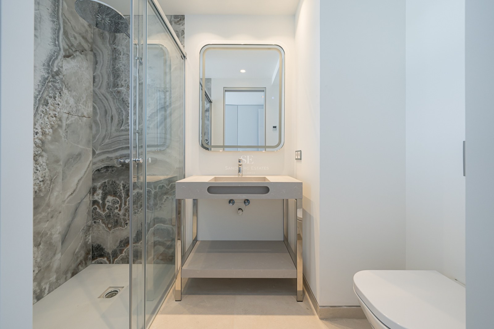 Baño moderno con ducha de cristal y paredes de mármol, espejo retroiluminado y lavabo minimalista.