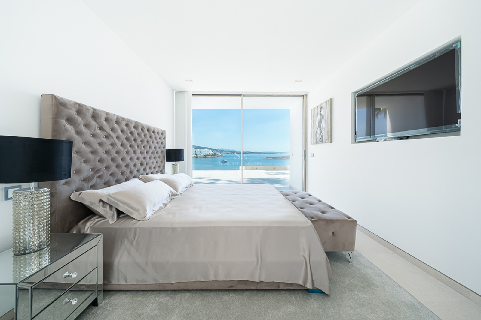 Dormitorio moderno con cabecero capitoné, muebles de espejo y grandes ventanales con vistas al océano azul.