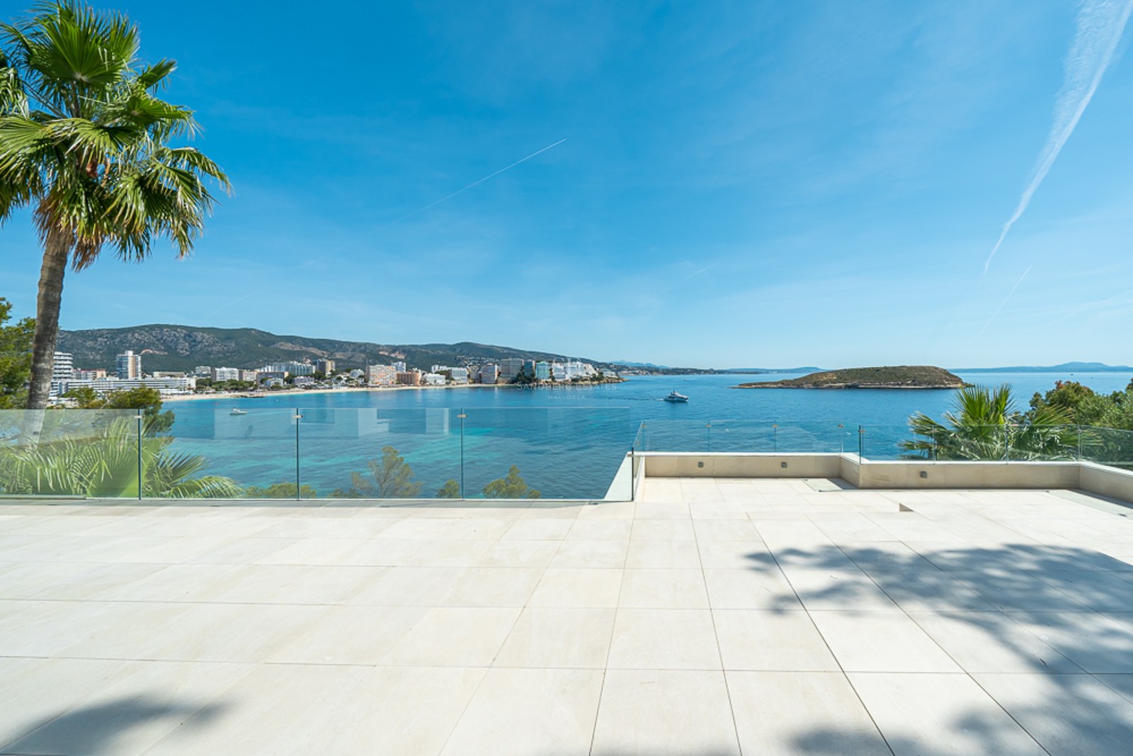 Amplia terraza de baldosas blancas con barandillas de cristal con vistas a una bahía azul y la costa.
